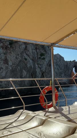 Boat deck with metal railing and orange lifebuoy passing steep rocky coastal cliffs. Maritime travel scene on a sunny day at sea.
