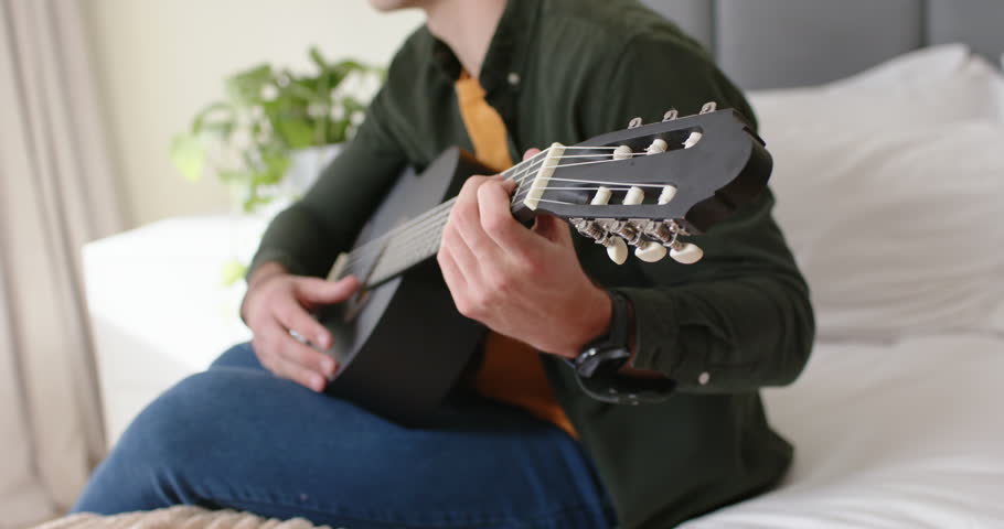 African American man settling cross-legged, playing black guitar on bed in bedroom, practicing song. Instrument, acoustic, fingerstyle, chords, strings, melody, solo