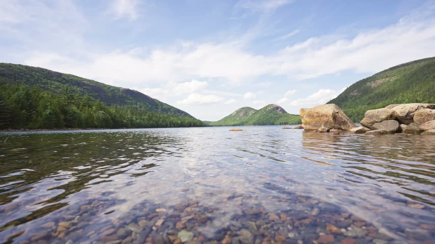 Clear waters and granite boulder-lined shore of Jordan Pond stretching toward distant forested mountains in Acadia National Park on Mount Desert Island, Maine.