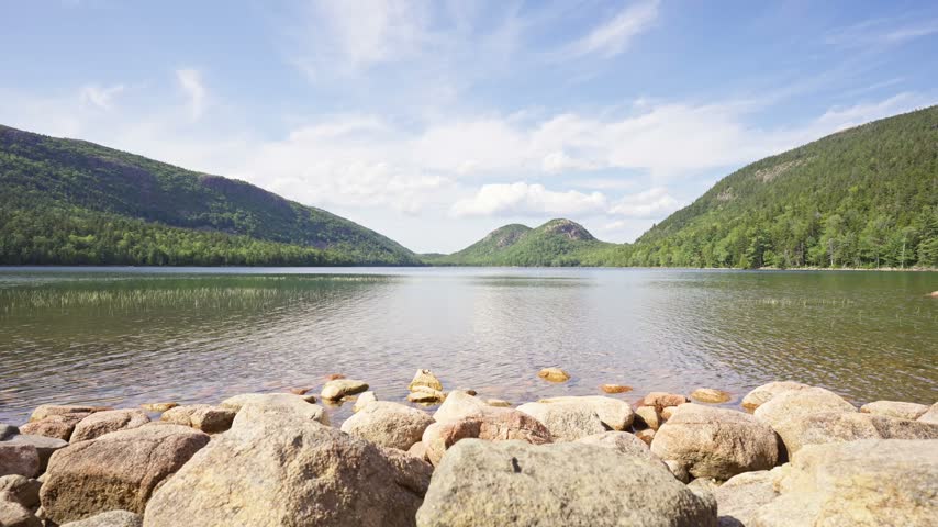 Clear waters and granite boulder-lined shore of Jordan Pond stretching toward distant forested mountains in Acadia National Park on Mount Desert Island, Maine.