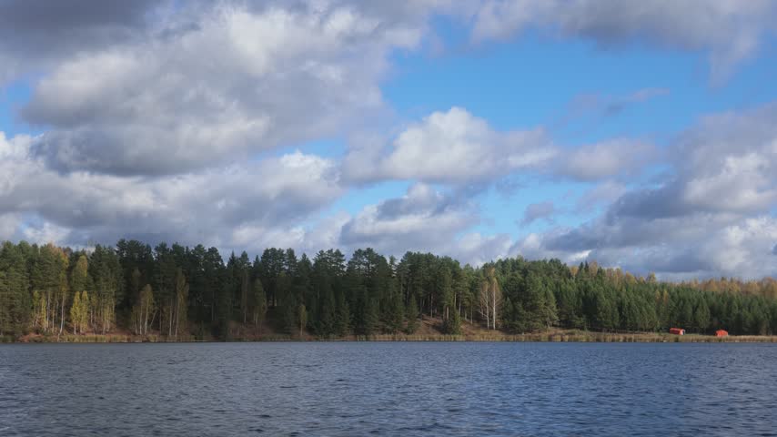 Scenic slow pan of an autumn camping site on a lake shore with tents and a colorful mixed forest under a cloudy sky.