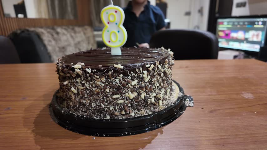 Chocolate cake covered in nuts sits on a table. Child holds a number eight candle. Person smiles behind the cake. Warm indoor lighting shows a celebration. Great for birthday or child events
