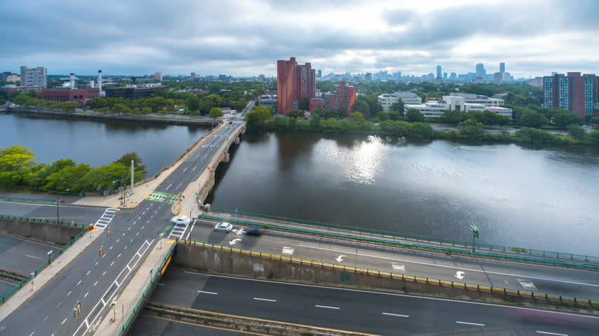 An elevated timelapse view of cars driving across a bridge spanning the Charles River, with the Boston skyline visible in the distance under a cloudy sky.