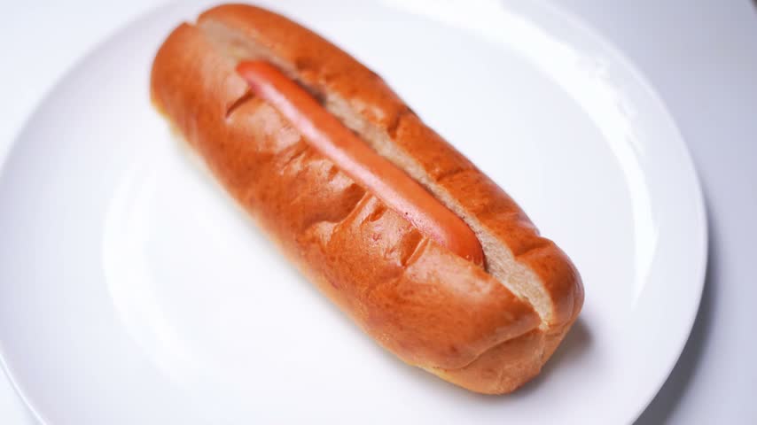 Hand Applying Creamy White Condiment in Wavy Lines on Fresh Hot Dog Bun.