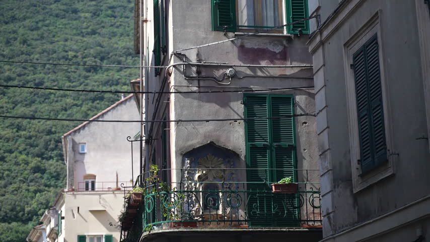 Historic stone buildings rise above mountain village, with green shutters and ornate balconies facing wooded slopes. This alpine town architecture offers picturesque setting for mountain tourism.