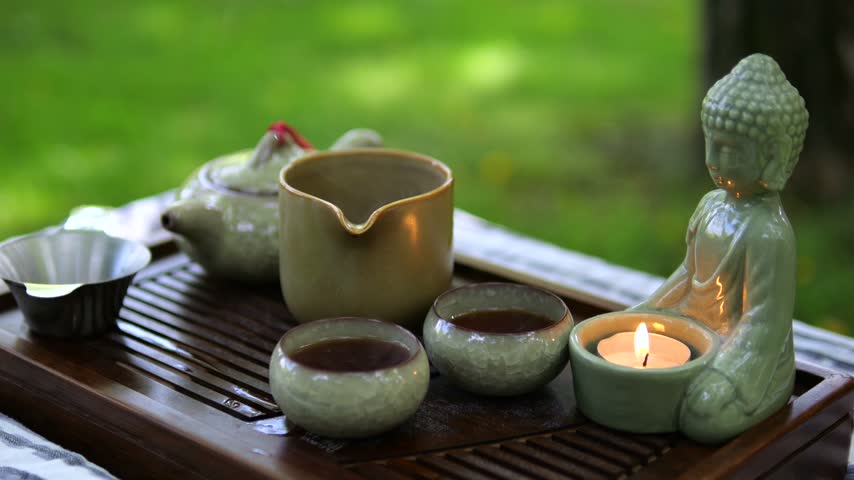 Glazed ceramic tea vessels surround glowing candle in serene garden arrangement. Buddha shaped holder and handcrafted bowls bring calming touch to outdoor tea and meditation, arranged with ceramic