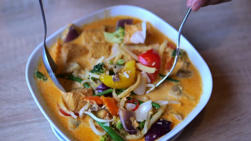 Diners mix vegetable curry in white bowl with spoons at restaurant table. Creamy orange sauce coats tofu and colorful vegetables as hands prepare this inviting meal to enjoy together.