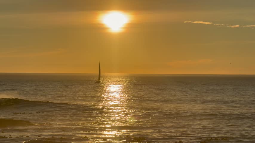 Silhouetted sailboat on calm ocean at golden sunset with sun glowing through clouds