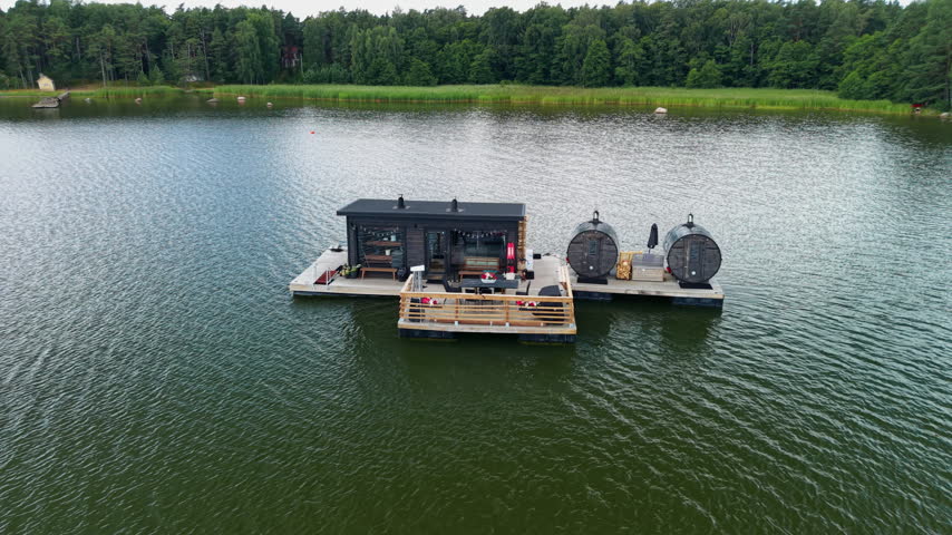 Floating sauna platform on a calm Finnish lake surrounded by dense green forest, featuring barrel saunas, wooden deck, and outdoor seating area