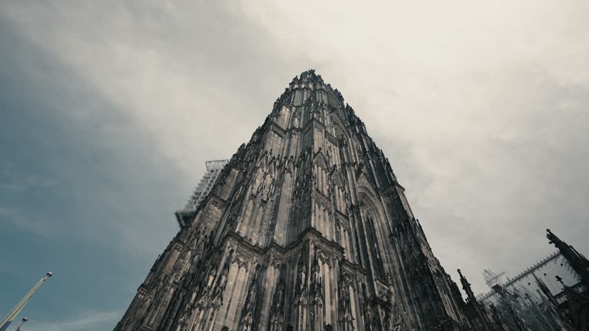 Cinematic low angle perspective looking up at the massive gothic tower of Cologne Cathedral in Germany. Features intricate dark stone architecture under a dramatic cloudy sky