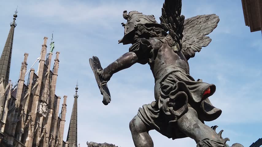 Ornate winged angel statue holding a sword with historic church spires and blue sky visible behind it in Munich Germany under bright daylight