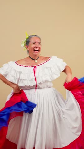 Graceful senior woman with gray hair wearing a vibrant, traditional folk costume, smiling and joyfully performing a latin american cultural dance against a plain studio background