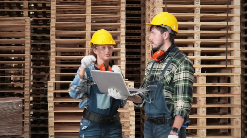 Two factory workers in safety helmets discuss production data on a laptop among stacked wood pallets inside a busy lumber warehouse.