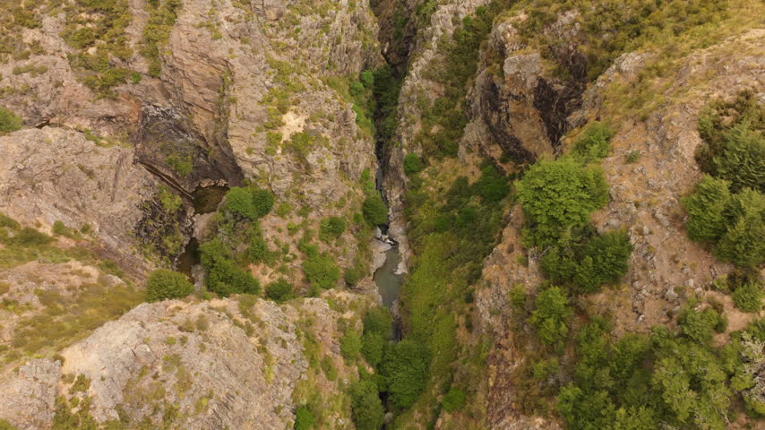 Wide aerial view drifting above the rocky Woolshed Creek gorge, revealing Emerald Pool, a narrow stream and scattered green vegetation near Mount Somers, New Zealand.