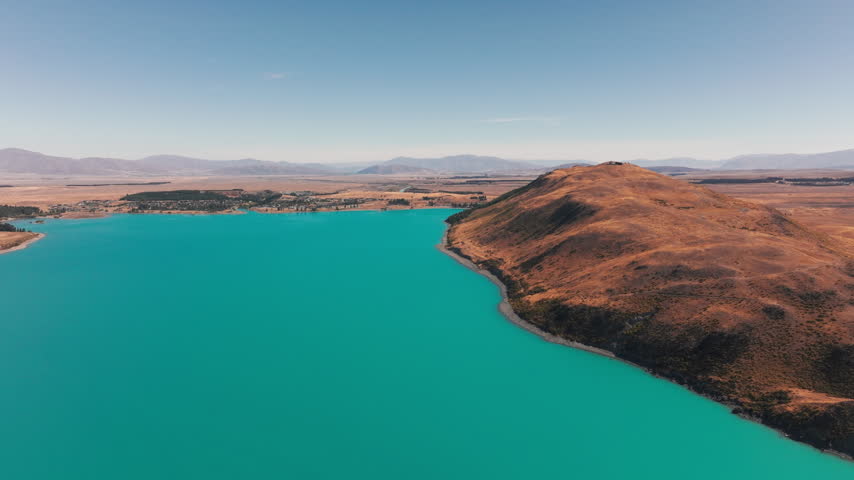 An aerial view follows the curved shore of Lake Tekapo beside a golden hillside. Turquoise water contrasts with dry grassland and distant mountains in New Zealand.