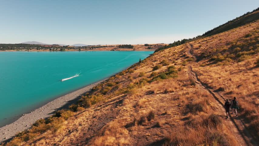 Two hikers follow a golden hillside trail while a motorboat leaves a white wake on Lake Tekapo. An aerial view combines lakeside walking and boating in New Zealand summer scenery.