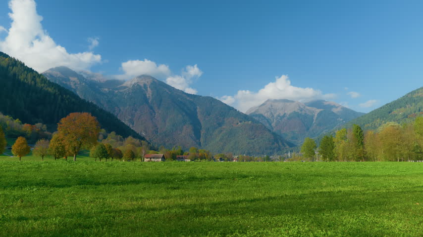 Mountain valley green meadow peaks under a blue sky with white clouds. Scenic alpine countryside village with autumn trees in a rural landscape, static shot.
