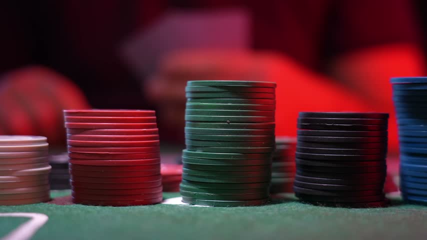 Stacked poker chips in red, green, black, and blue sit on green felt. The background is dark with red lighting. Casino atmosphere is clear. Scene is calm and professional