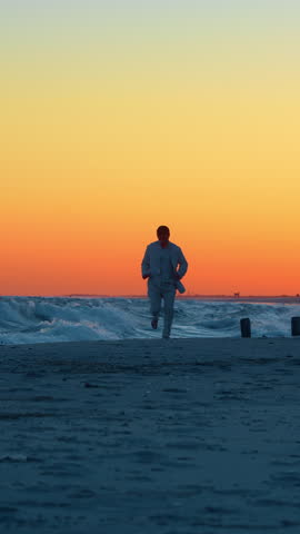 Man running on beach beside breaking waves at sunset. A barefoot man in a pale suit and dark cap moves toward the camera, with foaming water below an orange sky.