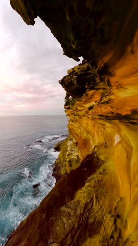 Slow motion vertical panning footage of golden sandstone cliffs and ocean waves at sunrise along the Sydney coastline, New South Wales, Australia.