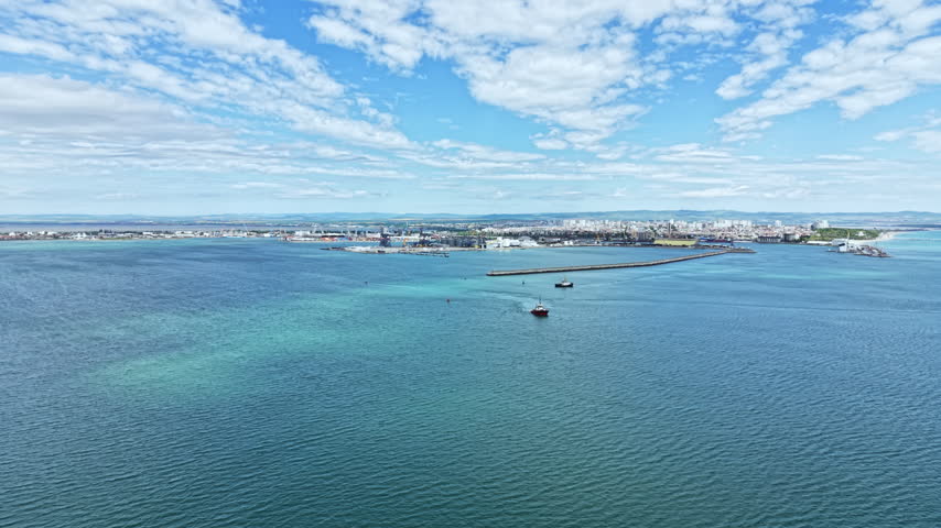 A wide view shows boats on the water near a harbor. The city skyline is visible in the background under a blue sky with clouds. It is late afternoon.