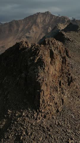 Dramatic aerial orbit around the rocky ridge of kumbeltau peak. Majestic snow-capped mountains and a cloudy sky in the background