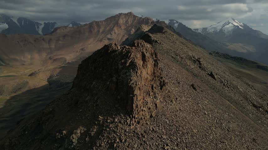 Dramatic aerial orbit around the rocky ridge of kumbeltau peak. Majestic snow-capped mountains and a cloudy sky in the background