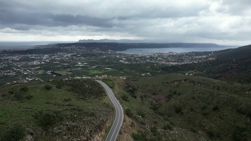 Aerial drone flight over green hills and a winding road near Chania, with a view across the plain and villages to Souda Bay under an overcast winter sky, Crete, Greece.
