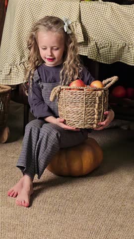 Smiling barefoot little girl in a gray outfit sitting on a pumpkin and holding a wicker basket of apples, table and pumpkins behind. Fall harvest concept, grainy film texture