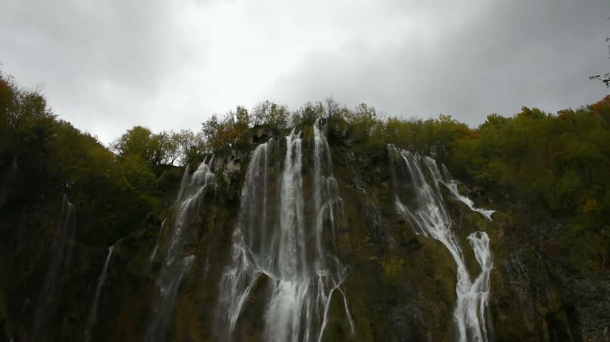 A stunning waterfall cascading powerfully from towering rocky cliffs draped in lush autumn foliage under a cloudy sky in Plitvice Lakes National Park, Croatia. The dynamic, vertical composition highlights the frothy water flow, creating a natural, wild, and serene atmosphere. Ideal for ecotourism projects, nature documentaries, and travel campaigns.
