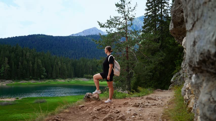 Hiker pausing on rocky forest trail overlooking tranquil green mountain lake, admiring scenic wilderness landscape and distant peaks under soft daylight sky