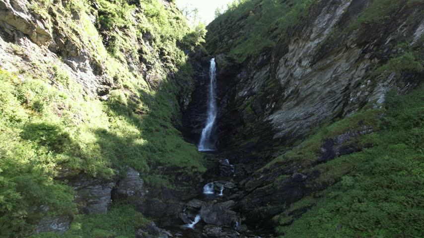 Tall thin waterfall in rocky gorge, slow aerial flight in Vestland, Norway, filmed on a sunny summer day.