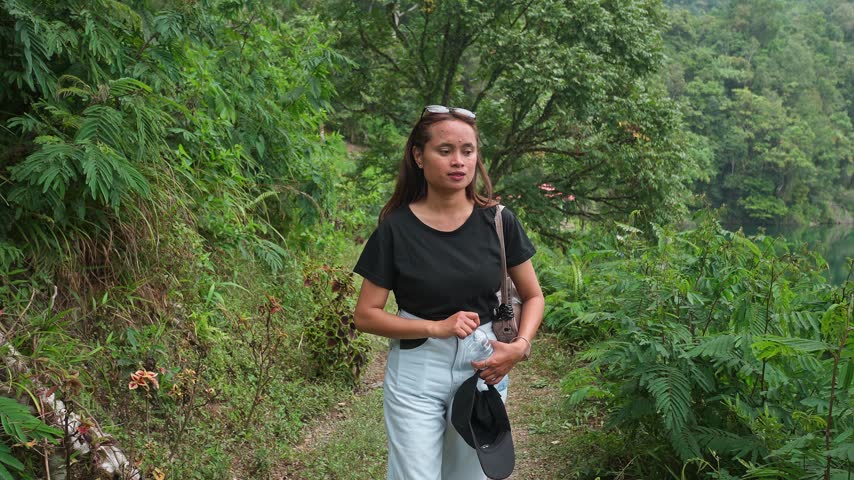 Indonesian woman walking forward on a trail through a green forest with a lake view.
