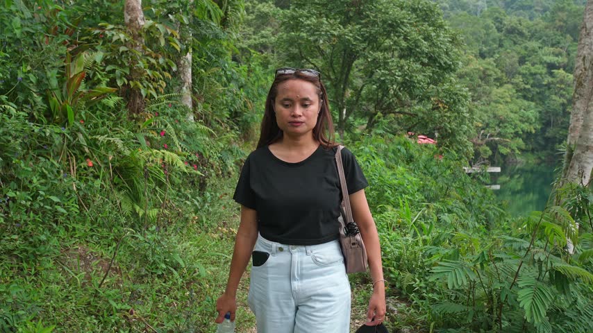 Indonesian woman walking forward on a trail through a green forest with a lake view.
