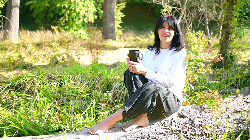 Thoughtful young brunette woman drinking coffee and sitting on a log slow motion