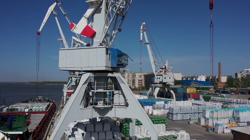 Large cargo cranes unload containers from a ship docked at a river port during daytime with clear blue skies and urban buildings in the background.