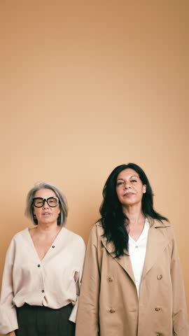 Two stylish and confident senior businesswomen posing back to back. An elegant grey haired lady and her brunette friend smiling at the camera. Vertical