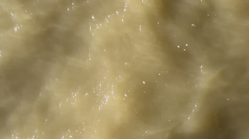 Top down view of fast moving muddy river water with bright sun reflections and turbulent current