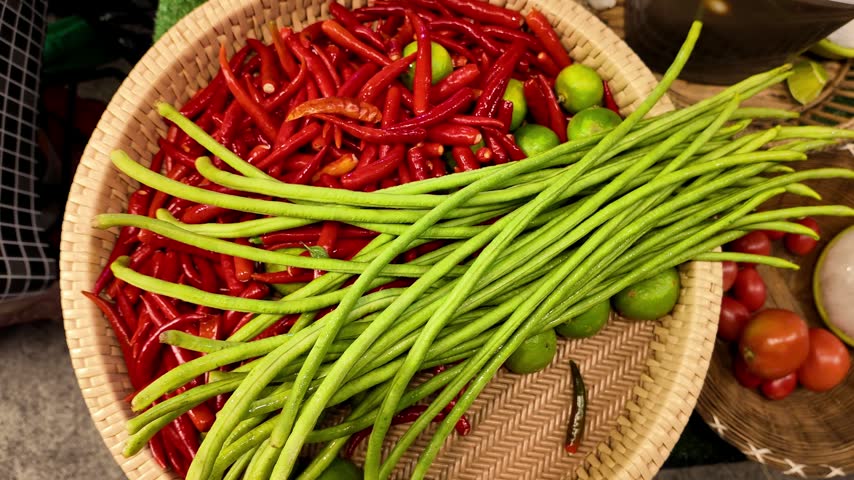 Close up shot of fresh raw ingredients including red chili peppers, long green beans and limes inside a wicker basket at a traditional market stall in Thailand.
