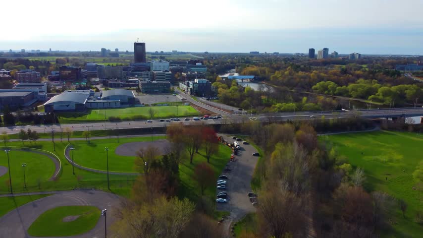 High-angle scenic aerial view looking south over the Carleton University campus in Ottawa, Ontario, Canada, showcasing university sports fields, multi-lane roadway corridors with commuter traffic, the winding path of the Rideau River, and sprawling parklands with early autumn tree foliage under a partly cloudy sky.