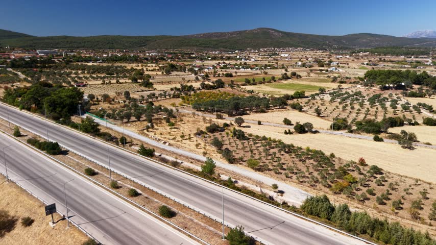 4K aerial drone footage of a divided highway running through olive groves and dry countryside toward forested hills.