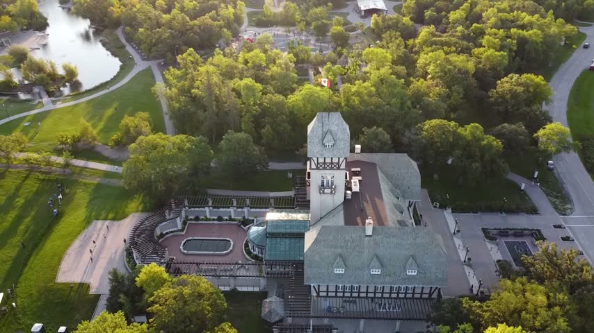 High-angle scenic aerial view looking over the historic architecture of the Pavilion at Assiniboine Park in Winnipeg, Manitoba, Canada, showcasing the iconic Tudor-style tower, a landscaped formal garden plaza, expansive green event lawns with long late-afternoon shadows, and the park