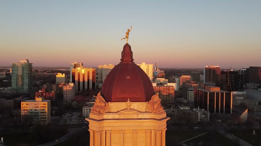 High-angle scenic aerial drone perspective focusing on the iconic gilded Golden Boy statue atop the Manitoba Legislative Building dome in Winnipeg, Manitoba, Canada, showcasing a sprawling background layout of downtown corporate skyscrapers, high-density brick apartments, and office towers bathed in a vibrant sunset golden hour glow with long dramatic shadows.