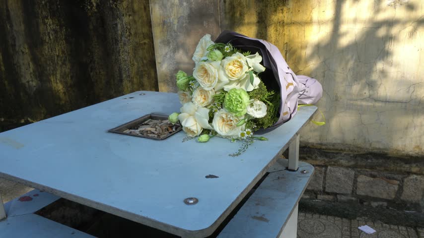 A wrapped bouquet of white roses rests on a table. The surrounding landscape is in Vietnam.