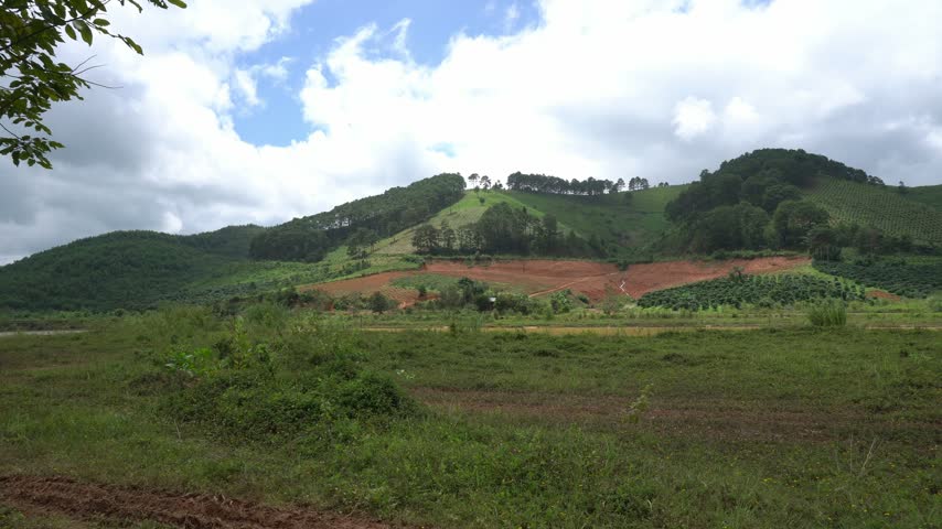 A rural hillside covered with green vegetation and patches of exposed red soil appears beneath clouds.