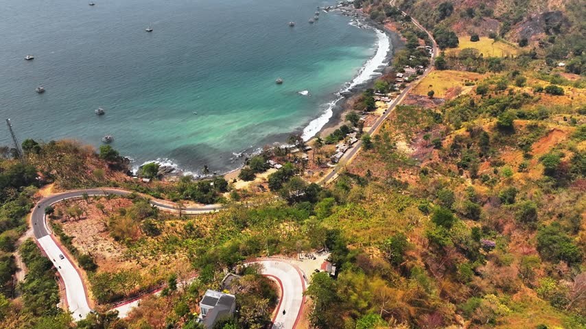 An overhead aerial perspective captures a dramatic coastal landscape featuring a winding concrete highway descending toward turquoise waters. Bright daylight illuminates lush rolling hillsides dotted with scattered buildings alongside a dark sand beach where gentle waves break. Several small vessels float peacefully across the expansive sea under clear skies during noon.