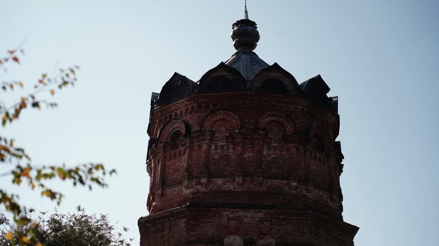 A weathered red brick tower with arched niches and a dark metal onion-shaped cupola rises against a pale blue sky, framed by branches with yellowing autumn leaves in the foreground.