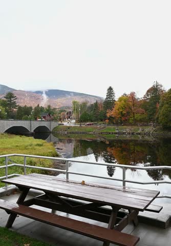 Wooden picnic table by river with stone bridge and autumn mountains behind