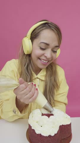 Cheerful female baker piping white frosting onto a red velvet cake against a pink background