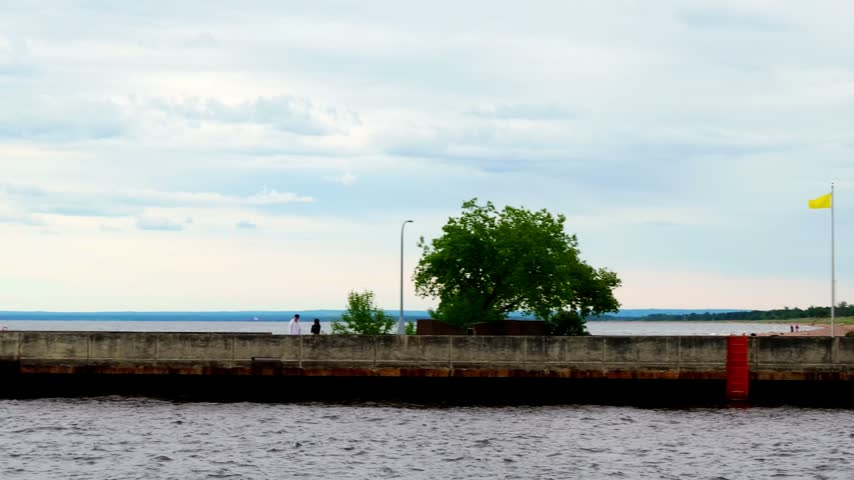 Tall black steel lighthouse tower with white lantern room and gallery railing stands near the Lake Superior shoreline on the Duluth South Breakwater, Minnesota; concrete seawall and yellow flag visible.